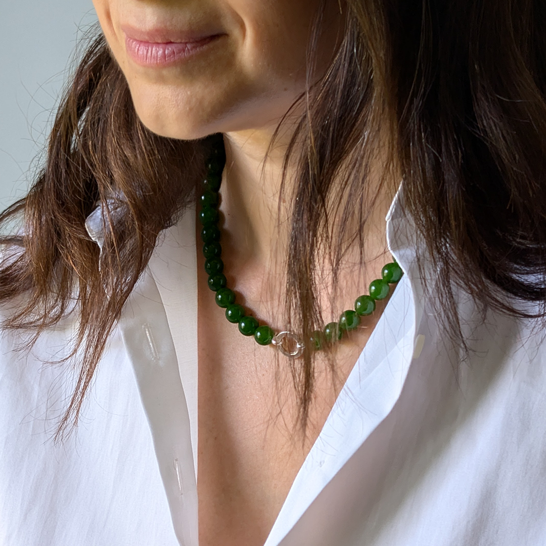 A close-up of a woman wearing a green jade beaded necklace with a clear round stone accent, styled with a white button-down shirt.