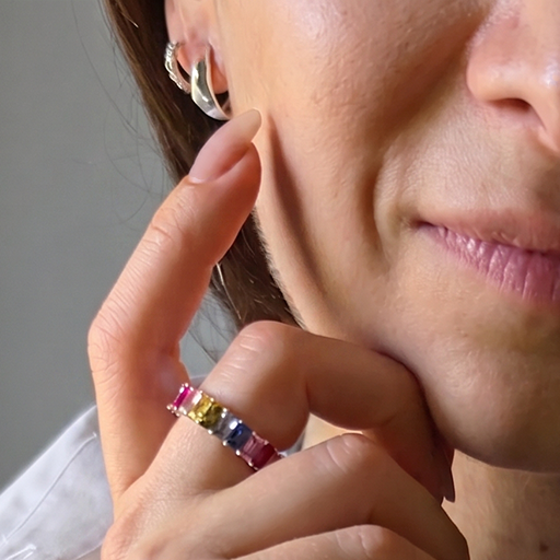 A close-up lifestyle shot of a woman wearing a light blue beaded necklace and a matching pink beaded bracelet, with a colorful ring on her finger, set against a neutral gray background.