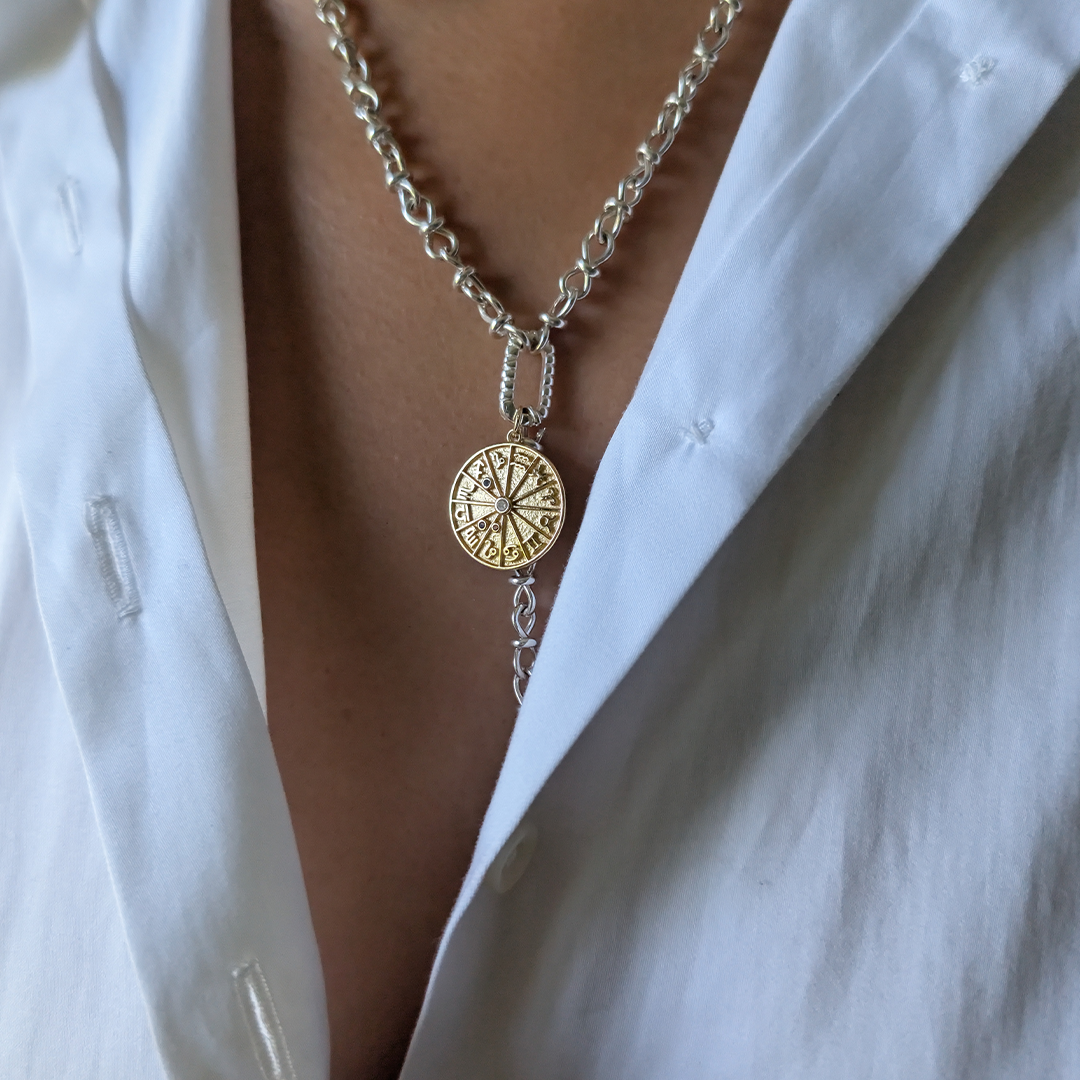 A close-up of a person wearing a silver chain necklace with a circular gold pendant featuring a zodiac design, styled over an open white shirt.